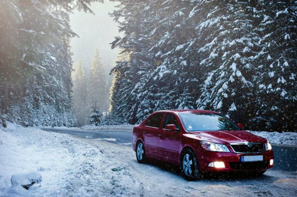 Essential Car Maintenance Tips 3 Red car driving on a snow-covered road surrounded by trees, emphasizing the need for winter car maintenance and safety