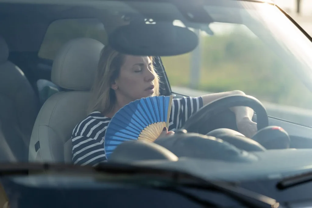 Frustrated woman fanning herself inside a hot car with broken AC