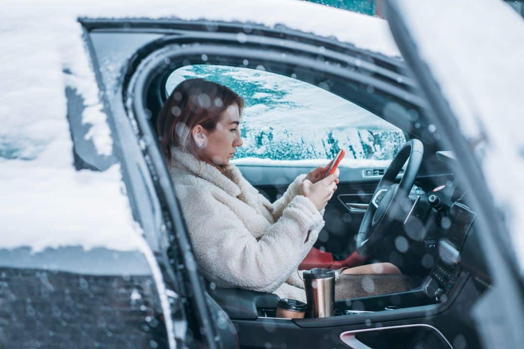 Woman driver checking phone inside car during winter — cooling system protection and car maintenance in Newark NJ