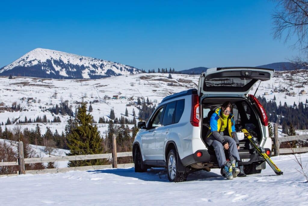Driver preparing winter gear next to an SUV parked in a snowy mountain area before a cold weather road trip